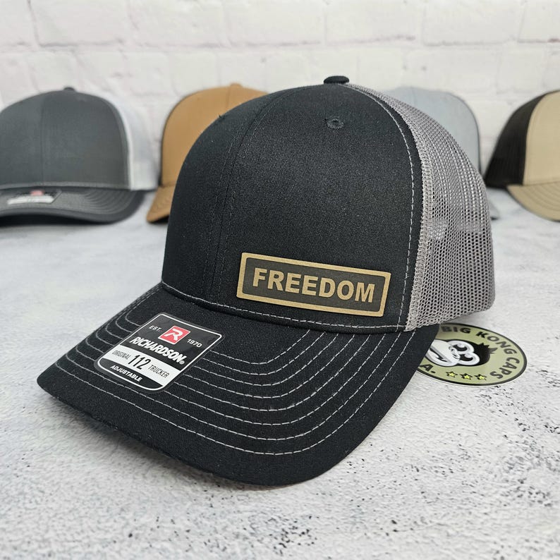 May include: Black trucker hat with a gray mesh back and a curved brim. The hat features a rectangular patch with the word "FREEDOM" in gold lettering. The hat has a Richardson 112 Trucker adjustable tag.