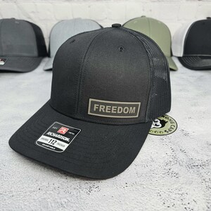 May include: Black trucker hat with a mesh back and a curved brim. The hat features a rectangular patch with the word "FREEDOM" in capital letters. The hat is adjustable and has a Richardson 112 tag.
