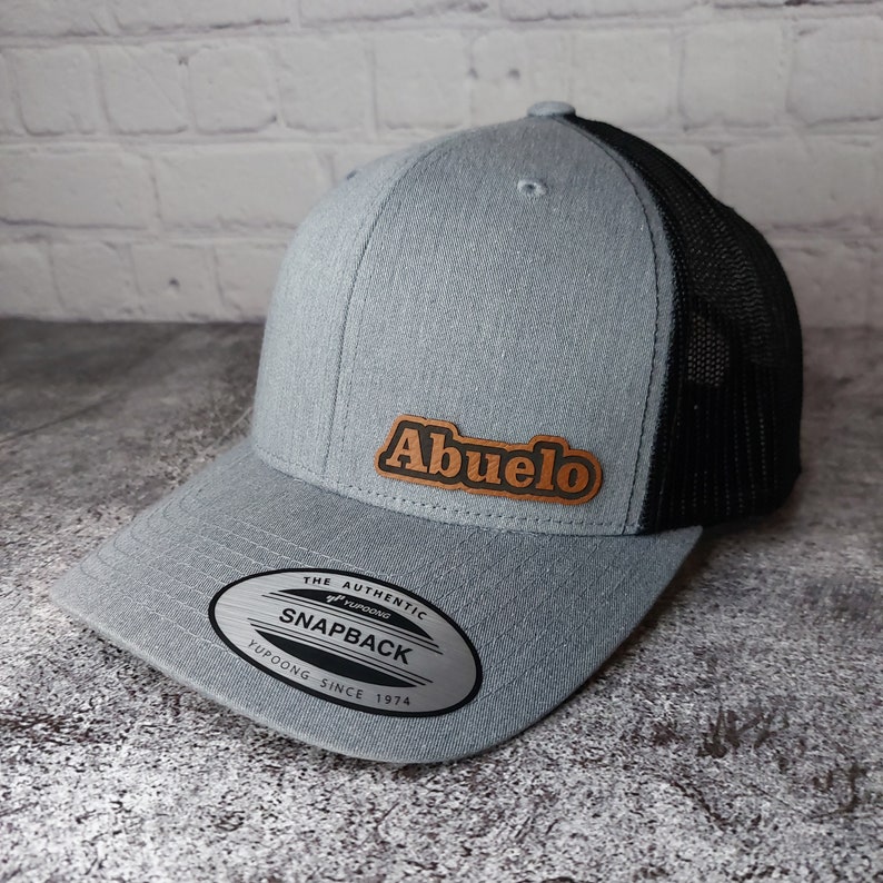 May include: A gray snapback hat with a black mesh back. The hat features a brown leather patch with the word "Abuelo" in orange lettering. The hat has a curved brim and a snap closure. The tag reads "The Authentic Snapback Yupoong Since 1974".