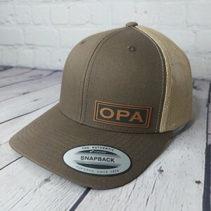 May include: A brown and tan baseball cap with a brown leather patch that says "OPA" on the front. The cap has a mesh back and a YUPOONG SNAPBACK label on the underside of the brim.