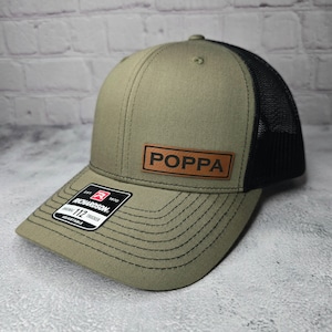 May include: Olive green and black trucker hat with a curved brim. The hat features a leather patch with the word "POPA" in capital letters. A black tag with the brand name "Richardson" is attached to the brim. The hat is adjustable.