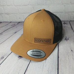 May include: A brown and black snapback hat with the word "GODFATHER" on a rectangular patch. The hat features a mesh back and a curved brim. The hat has a Yupoong Snapback label.