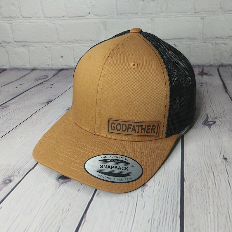 May include: A brown and black snapback hat with the word "GODFATHER" on a rectangular patch. The hat features a mesh back and a curved brim. The hat has a Yupoong Snapback label.