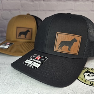 May include: Two baseball caps, one black and one tan, with a dog silhouette on a leather patch. The black cap has a black mesh back, and the tan cap has a tan mesh back. Both caps have a Richardson 112 Trucker tag.