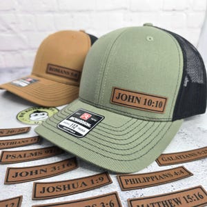 May include: Two trucker hats with leather patches. One is tan with "ROMANS 5:5" and the other is olive green with "JOHN 10:10". Additional patches with Bible verses are scattered around.