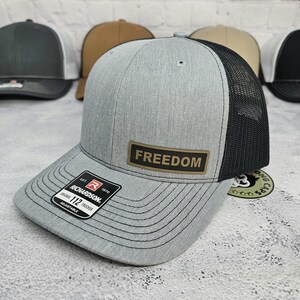 May include: A gray trucker hat with a black mesh back and a curved brim. The hat features a rectangular patch with the word "FREEDOM" and a Richardson patch. Adjustable fit.