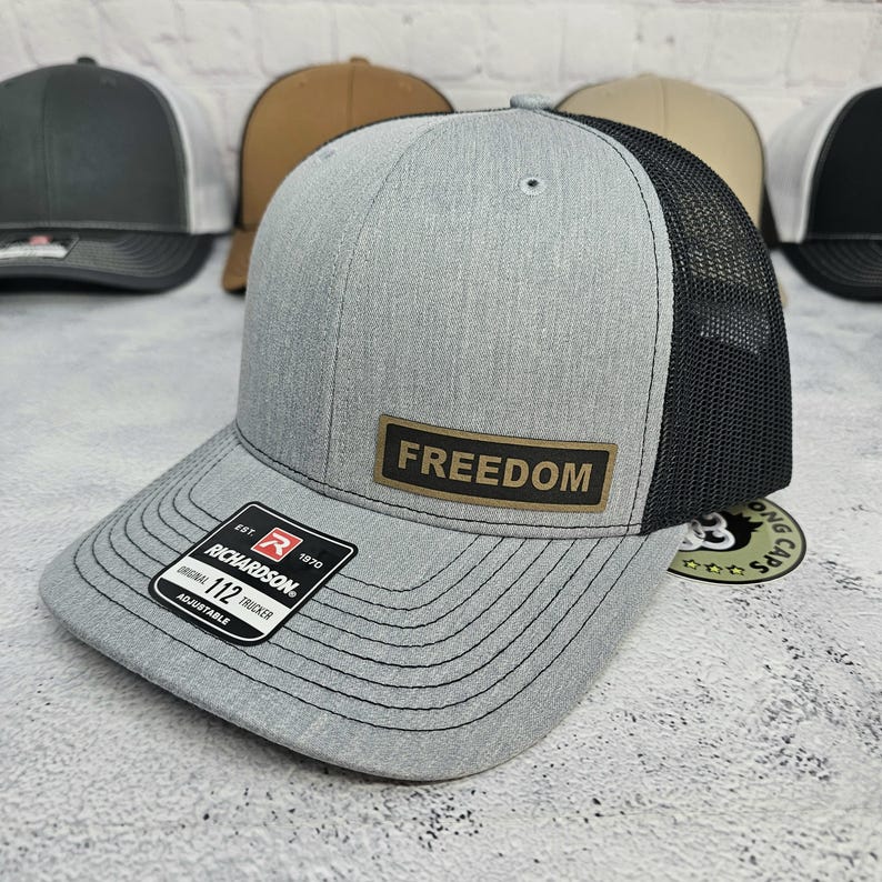 May include: A gray trucker hat with a black mesh back and a curved brim. The hat features a rectangular patch with the word "FREEDOM" and a Richardson patch. Adjustable fit.