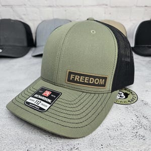 May include: Olive green trucker hat with a black mesh back and a curved brim. The hat features a rectangular patch with the word "FREEDOM" and a Richardson 112 adjustable tag. Several other hats are visible in the background.
