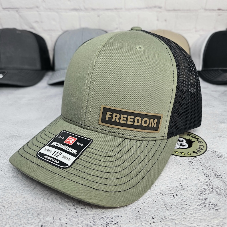 May include: Olive green trucker hat with a black mesh back and a curved brim. The hat features a rectangular patch with the word "FREEDOM" and a Richardson 112 adjustable tag. Several other hats are visible in the background.