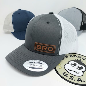 May include: A gray and white trucker hat with a brown leather patch that says "BRO" on it. The hat has a black mesh back and a gray bill. The hat is sitting on a white surface with a sticker that says "Big Kong U.S.A."