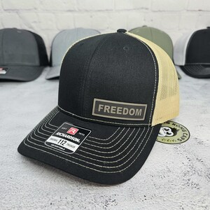 May include: Black and tan trucker hat with the word "FREEDOM" on a rectangular patch. The hat has a curved brim with white stitching and an adjustable strap. The Richardson brand label is visible.