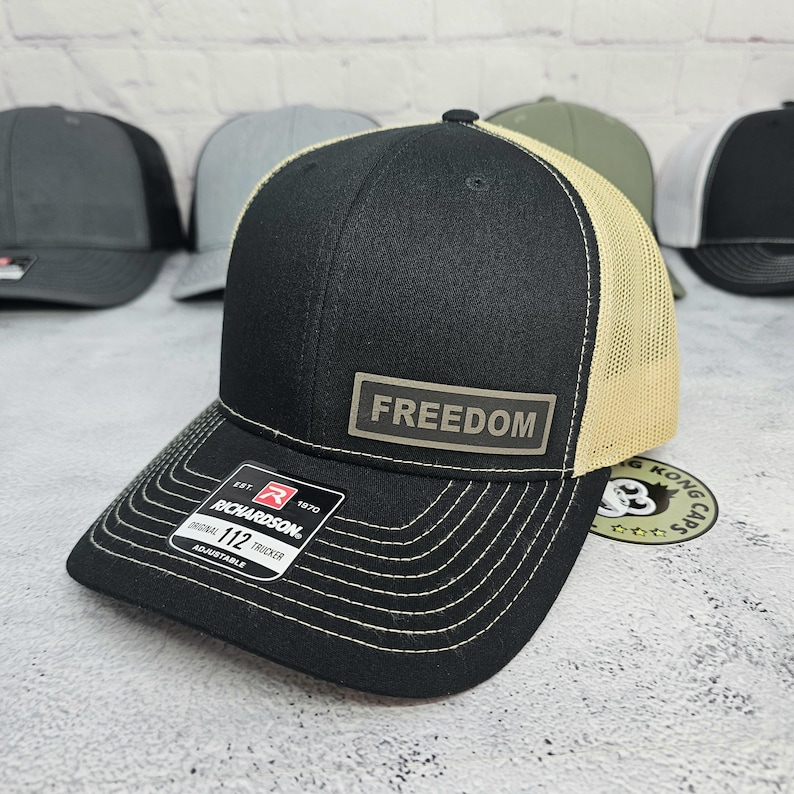 May include: Black and tan trucker hat with the word "FREEDOM" on a rectangular patch. The hat has a curved brim with white stitching and an adjustable strap. The Richardson brand label is visible.
