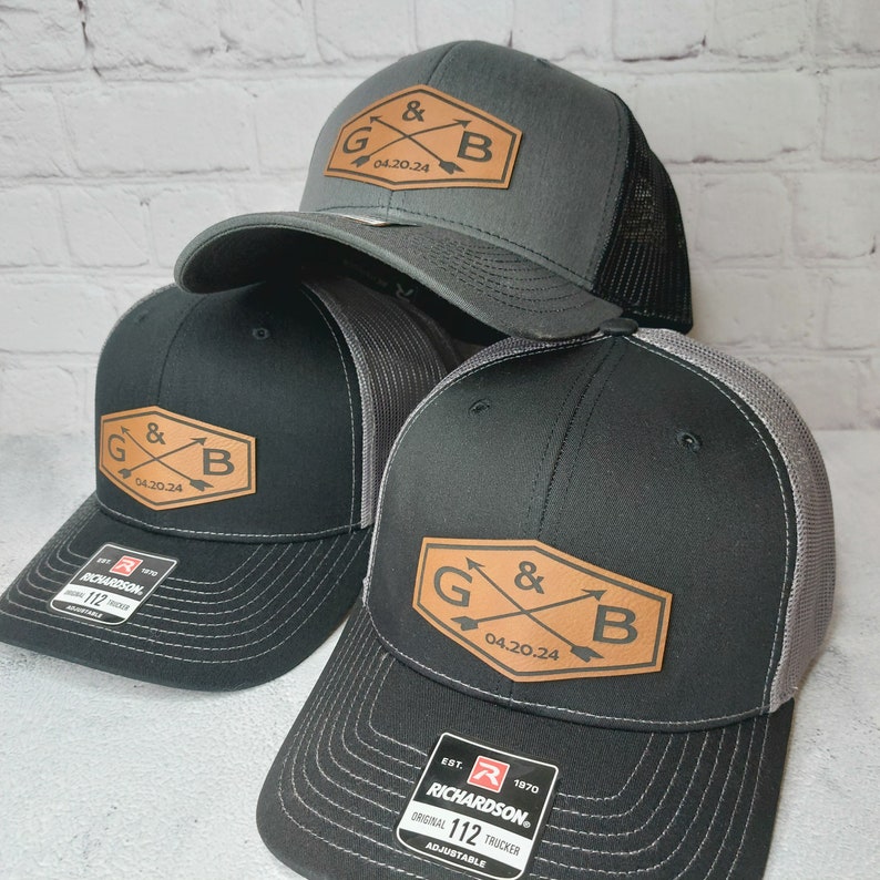 May include: Three black and gray trucker hats with a brown leather patch featuring the letters G and B with arrows crossing each other. The patch also includes the date 04.20.24. The hats are all adjustable and have a Richardson logo on the back.