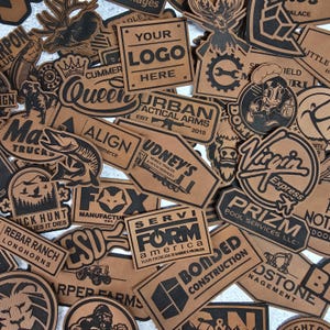 May include: A collection of brown leather patches with various designs and text. Some patches feature logos, while others display text such as "Your Logo Here", "Urban Tactical Arms", and "Bonded Construction". The patches vary in shape and size.