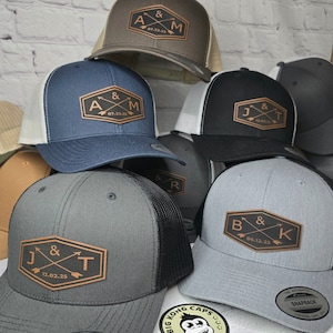 May include: Six different baseball caps with a brown leather patch on the front. Each patch has two arrows crossing each other and initials with a date. The caps are in different colors, including gray, black, brown, and blue.