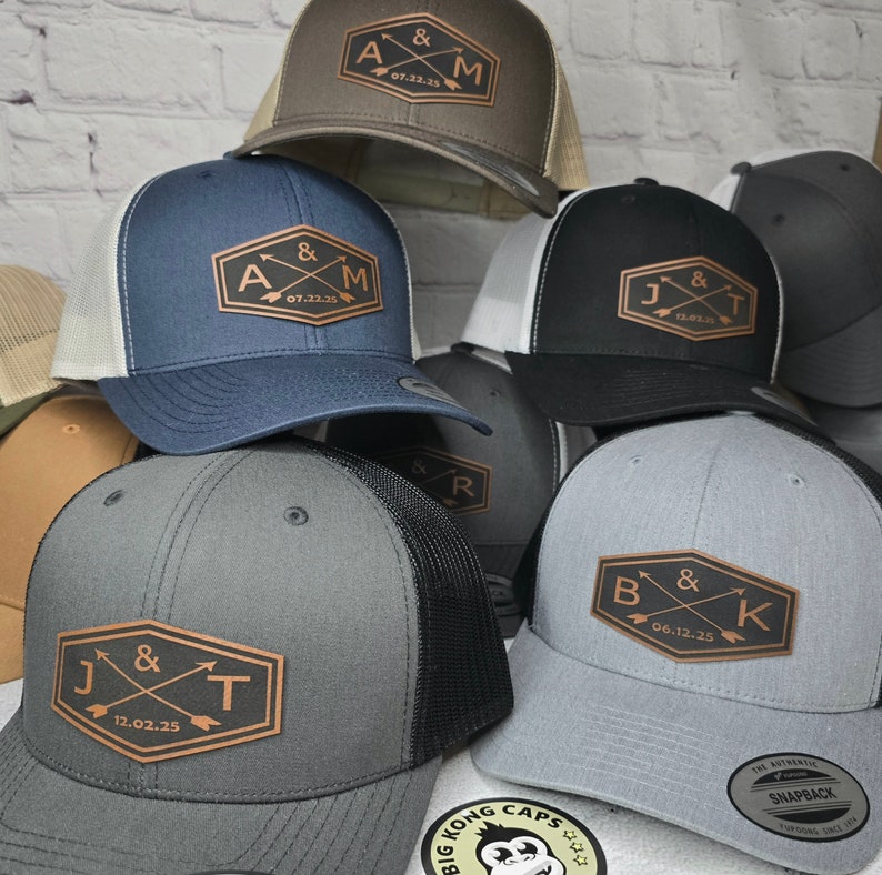 May include: Six different baseball caps with a brown leather patch on the front. Each patch has two arrows crossing each other and initials with a date. The caps are in different colors, including gray, black, brown, and blue.