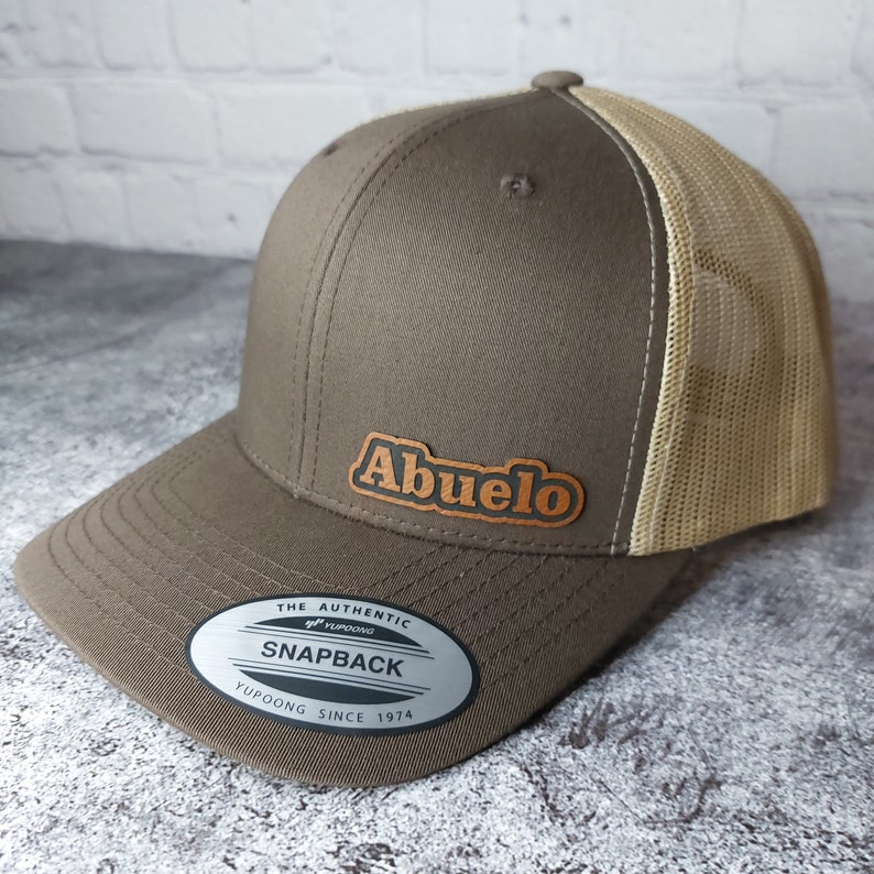 May include: A brown snapback baseball cap with a tan mesh back. The cap features the word "Abuelo" in a brown, stylized font on a leather patch. The hat has a curved brim and a snap closure.