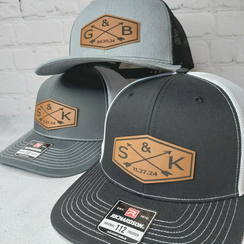 May include: Three trucker hats with leather patches featuring initials and dates. The hats are gray, black, and gray with white mesh. The patches are brown with white stitching.