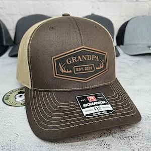 May include: A brown trucker hat with a tan mesh back and a curved brim. The hat features a brown leather patch with the word "GRANDPA" and "EST. 2026" printed on it. The hat is adjustable.