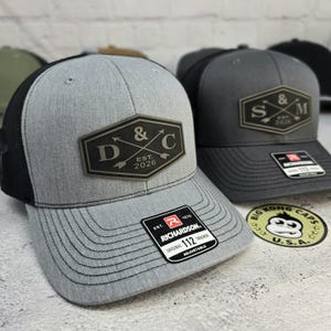 May include: Gray trucker hat with a black mesh back and a gray brim. The hat features a black and gray patch with the text "D & C EST. 2026". A Richardson 112 Trucker tag is visible. A Big Kong Caps sticker is also visible.