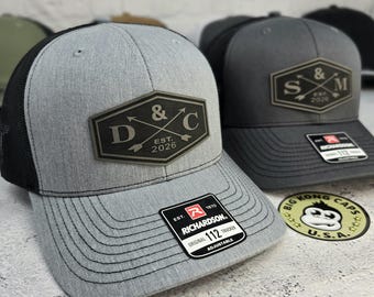 May include: Gray trucker hat with a black mesh back and a gray brim. The hat features a black and gray patch with the text "D & C EST. 2026". A Richardson 112 Trucker tag is visible. A Big Kong Caps sticker is also visible.