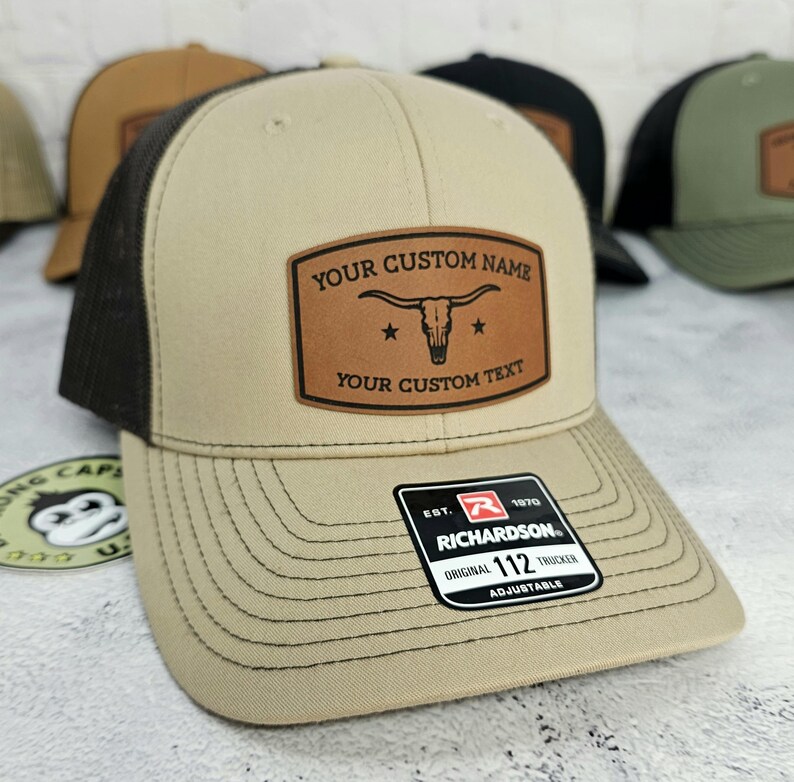 May include: Tan and black trucker hat with a leather patch. The patch features a longhorn skull and the text "YOUR CUSTOM NAME" and "YOUR CUSTOM TEXT". The hat has a curved brim and an adjustable strap. The tag reads "RICHARDSON 112 TRUCKER".