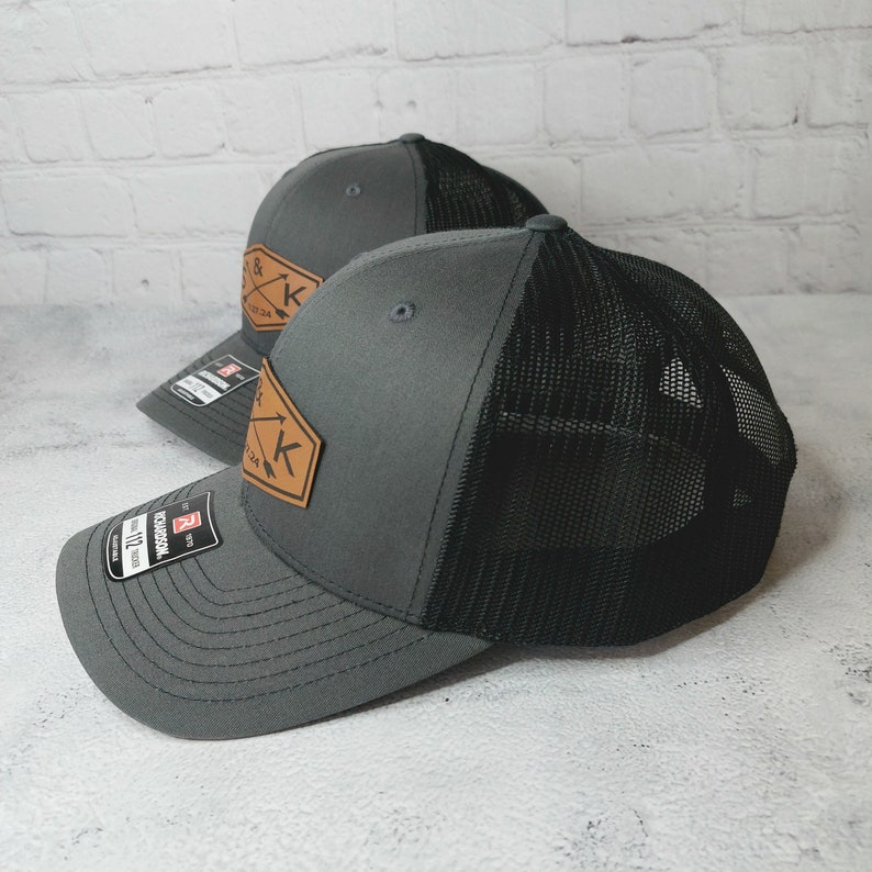 May include: Two gray and black mesh trucker hats with a brown leather patch featuring the letters "K & K" and arrows. The hats have a black mesh back and a gray front.