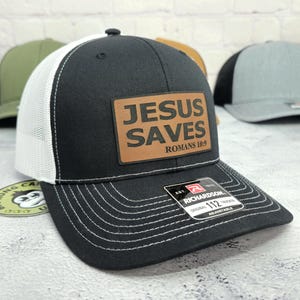 May include: Black trucker hat with a white mesh back and a curved brim. The hat features a brown leather patch with the words "JESUS SAVES ROMANS 10:9" in black lettering. The hat is adjustable and has a Richardson tag.