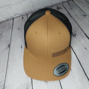 May include: A brown and black trucker hat with the word "GODFATHER" printed on the front. The hat has a mesh back and a snapback closure. The hat is made of a durable material and is suitable for everyday wear.