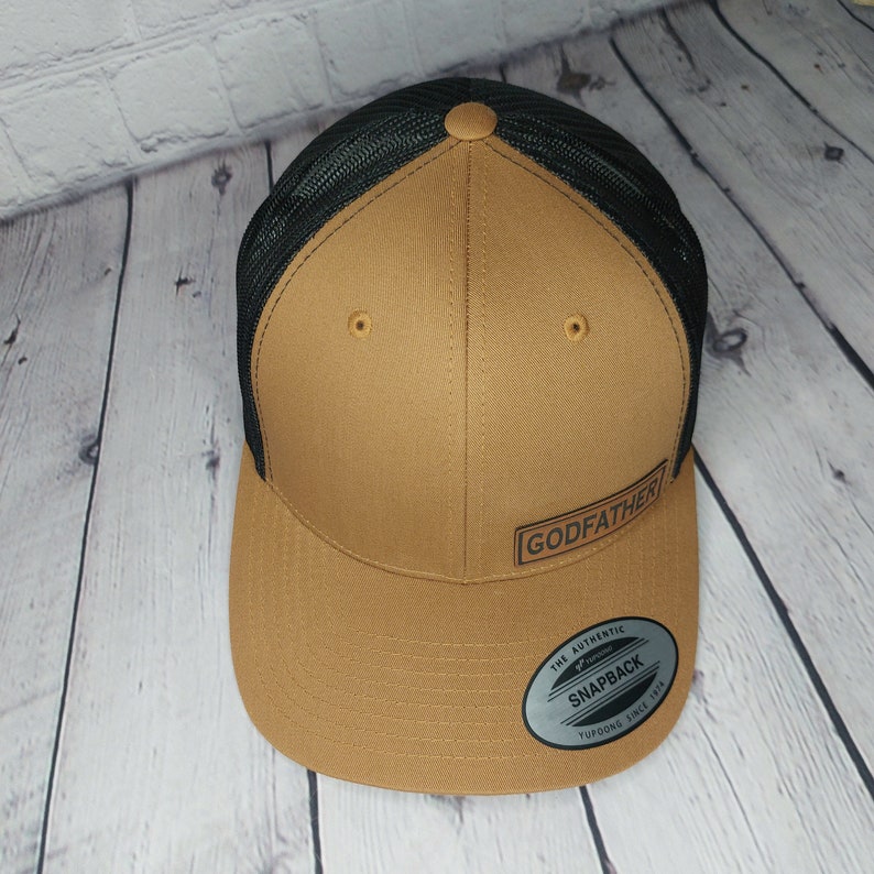 May include: A brown and black trucker hat with the word "GODFATHER" printed on the front. The hat has a mesh back and a snapback closure. The hat is made of a durable material and is suitable for everyday wear.
