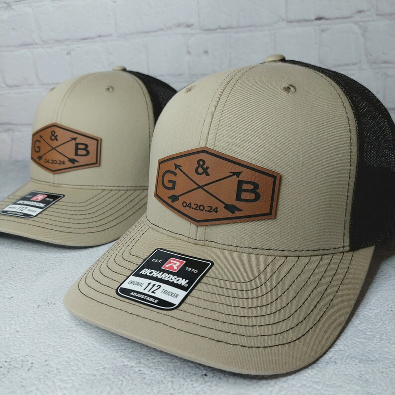 May include: Two beige and black trucker hats with a brown leather patch that says "G & B 04.20.24" in a diamond shape. The hats are adjustable and have a black mesh back.