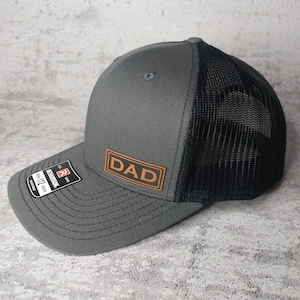 May include: A gray and black trucker hat with a brown leather patch that says "DAD" on it.