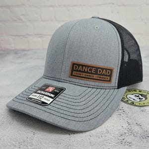 May include: A gray trucker hat with a black mesh back. The hat features a brown leather patch with the text "DANCE DAD I DON'T DANCE - I FINANCE". The hat is adjustable and has a curved brim.