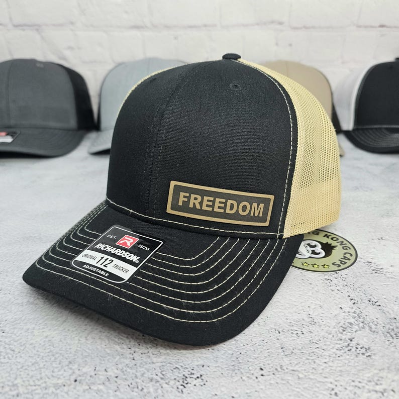 May include: Black and tan trucker hat with the word "FREEDOM" on a rectangular patch. The hat has a curved brim with white stitching and a mesh back. The brand is Richardson.