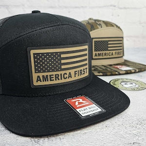 May include: Black flat bill snapback hat with a mesh back and a tan patch featuring an American flag and the words "AMERICA FIRST." A second hat with a camouflage pattern is visible in the background.