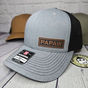 May include: A gray Richardson 112 Trucker hat with a black mesh back and a brown leather patch that reads "PAPAW." The hat has a curved brim and an adjustable strap. Additional hats are visible in the background.