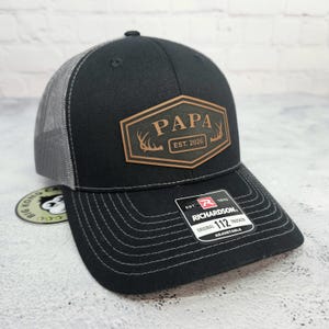 May include: Black trucker hat with a gray mesh back and a curved brim. The front features a brown leather patch with the word "PAPA" and "EST. 2026" along with deer antlers. The hat is adjustable.
