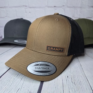 May include: A brown and black baseball cap with a black mesh back. The cap has a brown leather patch on the front that says "GRAMPY". The cap also has a silver sticker on the side that says "THE AUTHENTIC YUPOONG SNAPBACK YUPOONG SINCE 1974".