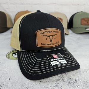 May include: Black and tan trucker hat with a leather patch that reads "VANSTARK RANCH FORTWORTH, TEXAS". The hat has a curved brim and a mesh back. The hat is adjustable and has white stitching.
