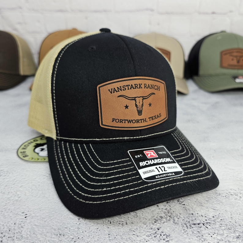 May include: Black and tan trucker hat with a leather patch that reads "VANSTARK RANCH FORTWORTH, TEXAS". The hat has a curved brim and a mesh back. The hat is adjustable and has white stitching.
