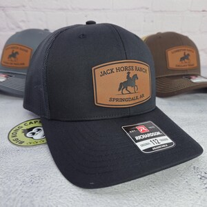 May include: Black trucker hat with a mesh back and a leather patch. The patch reads "Jack Horse Ranch Springdale, AR" with a silhouette of a person riding a horse. The hat is adjustable and has a Richardson tag.