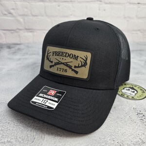 May include: Black trucker hat with a mesh back and a curved brim. The front features a tan patch with the word "FREEDOM" above crossed rifles and the year "1776". The hat has an adjustable strap and a "Richardson" tag.