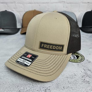 May include: Tan and black trucker hat with the word "FREEDOM" on a rectangular patch. The hat has a curved brim and a mesh back. The tag reads "RICHARDSON 112 TRUCKER ADJUSTABLE".