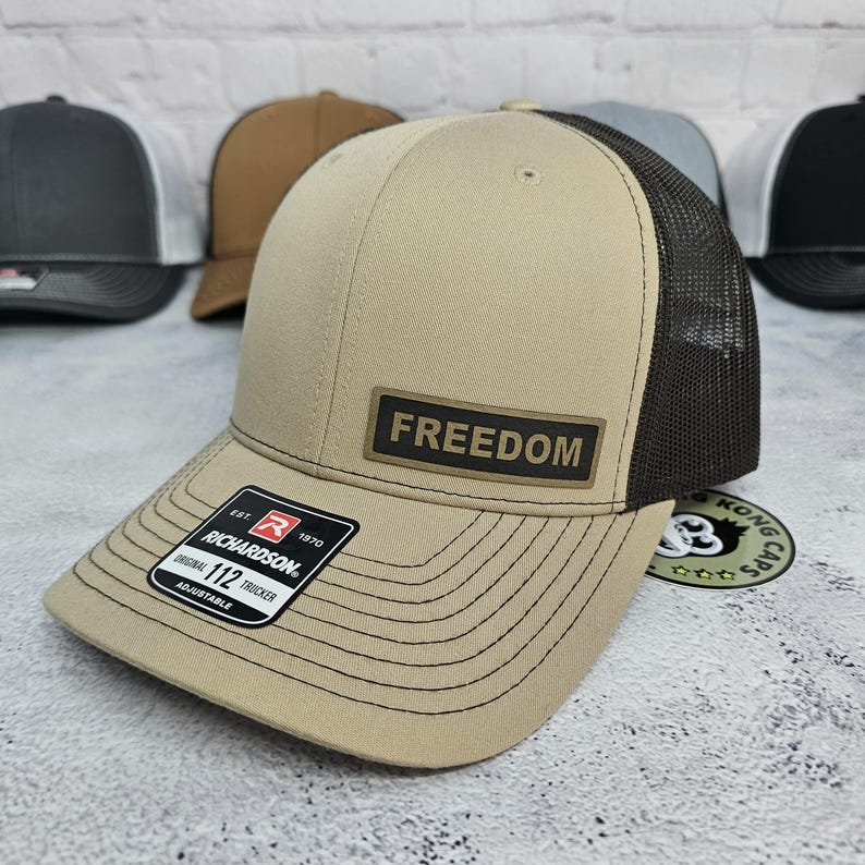 May include: Tan and black trucker hat with the word "FREEDOM" on a rectangular patch. The hat has a curved brim and a mesh back. The tag reads "RICHARDSON 112 TRUCKER ADJUSTABLE".