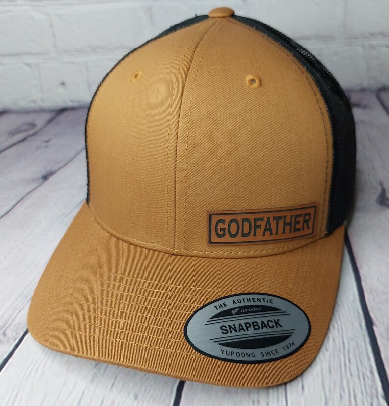 May include: A tan and black snapback hat with the word "GODFATHER" on a rectangular patch. The hat features a mesh back and a curved brim. The hat also has a "Yupoong" tag.