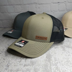 May include: A khaki-colored trucker hat with a black mesh back and a curved brim. The hat features a leather patch that reads "GRANDPA". The hat has a Richardson 112 adjustable tag.