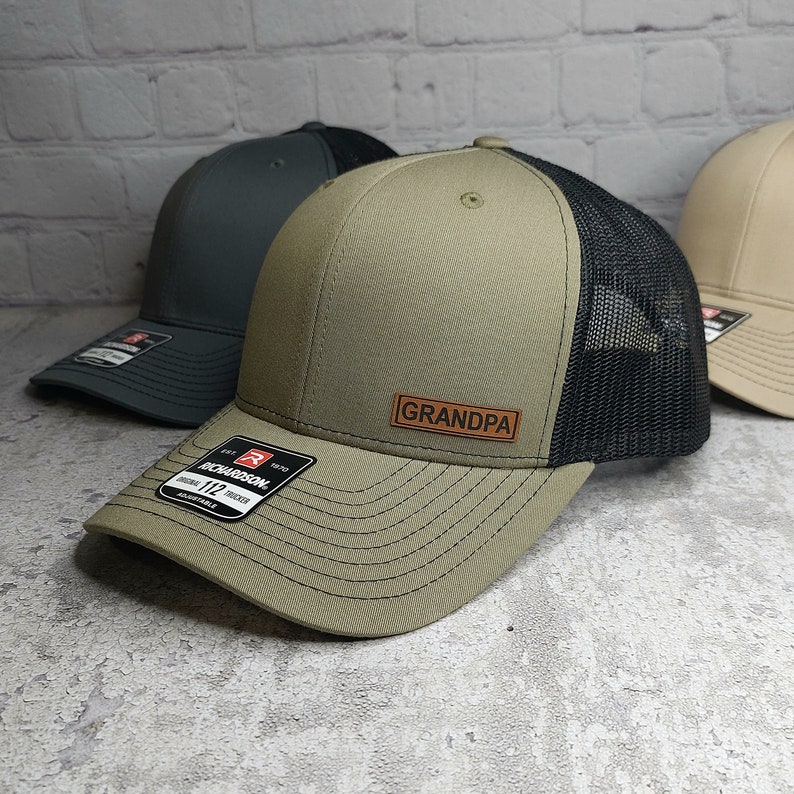 May include: A khaki-colored trucker hat with a black mesh back and a curved brim. The hat features a leather patch that reads "GRANDPA". The hat has a Richardson 112 adjustable tag.