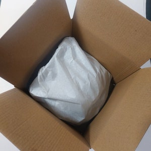 May include: A brown cardboard box with the top flaps open, revealing a white tissue paper-wrapped item inside. The box is empty except for the wrapped item, which is centered in the box.
