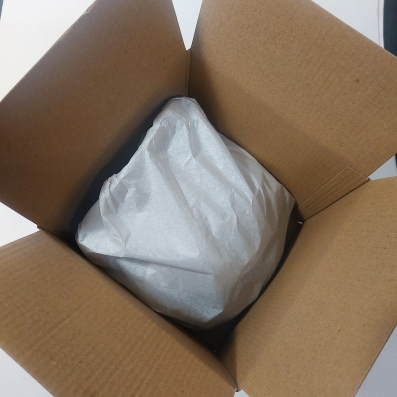 May include: A brown cardboard box with the top flaps open, revealing a white tissue paper-wrapped item inside. The box is empty except for the wrapped item, which is centered in the box.