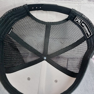 May include: A close-up view of the underside of a black and gray trucker hat. The hat features a black mesh back, a gray front panel, and a black adjustable snap closure. The brand name "RICHARDSON" is visible on a tag.